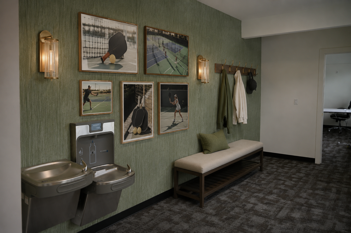 A wall with green wallpaper features four framed pickleball photos, a water fountain, a bench with a pillow, and a coat rack with hanging items.