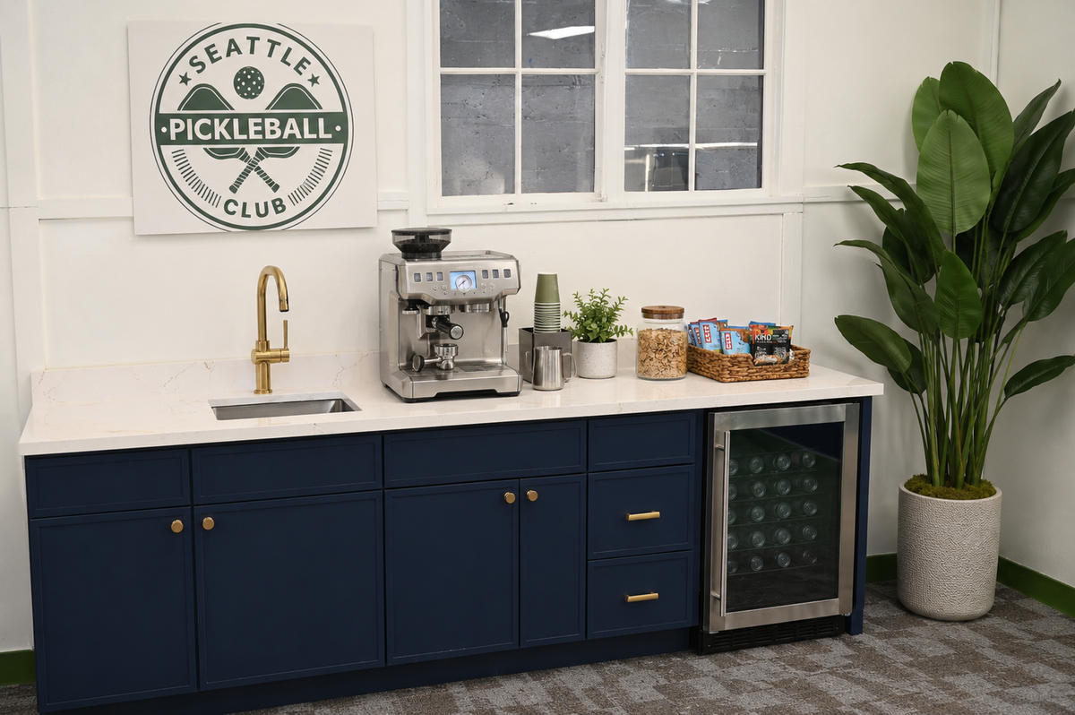 A refreshment station featuring a Seattle Pickleball Club sign, espresso machine, sink, snack basket, and under-counter beverage fridge.